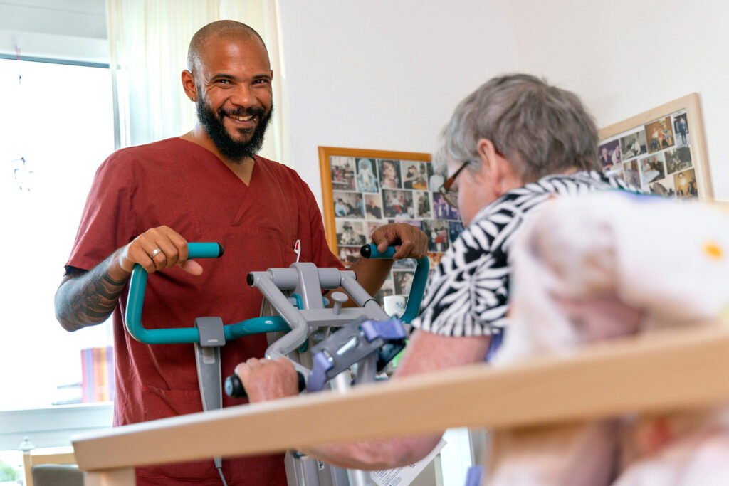 Ein Mitarbeiter unterstützt eine Bewohnerin beim Aufstehen und lächelt in die Kamera.