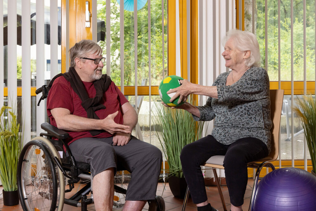 Ein Bewohner und eine Bewohner überreichen sich bei der Sitzgymnastik einen Ball.