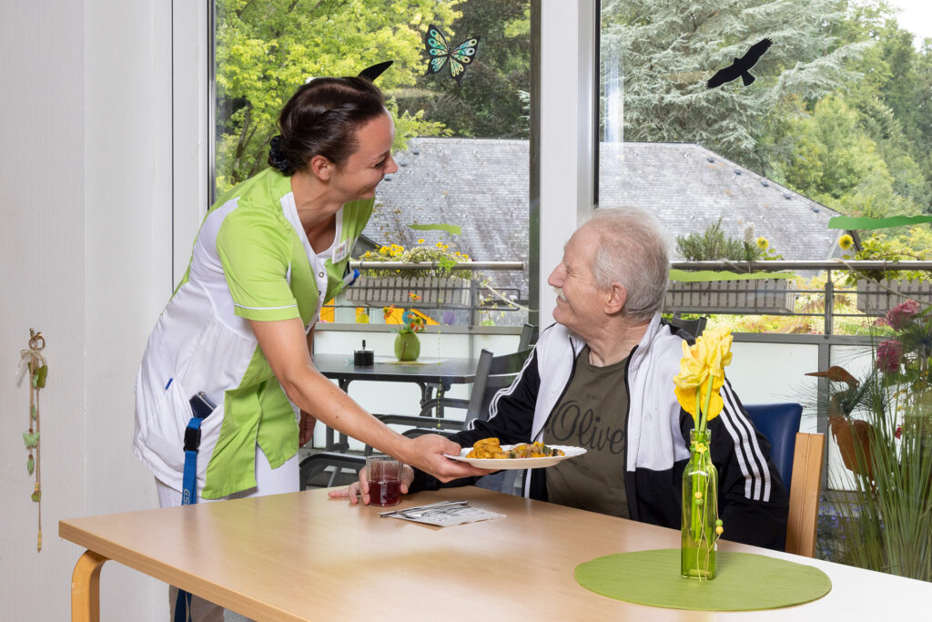 Eine Mitarbeiterin serviert einem Bewohner das Mittagessen am Tisch.