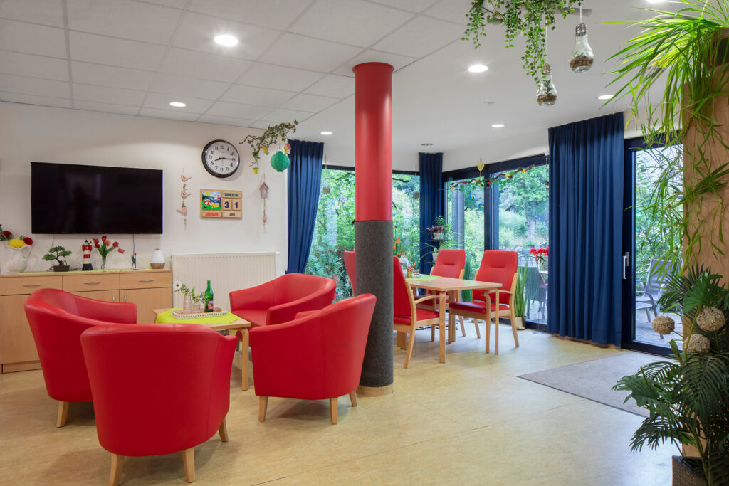Blick in einen hellen Aufenthaltsraum: Im Vordergrund stehen rote gemütliche Sessel, im Hintergrund eine Tischgruppe mit vier Stühlen.