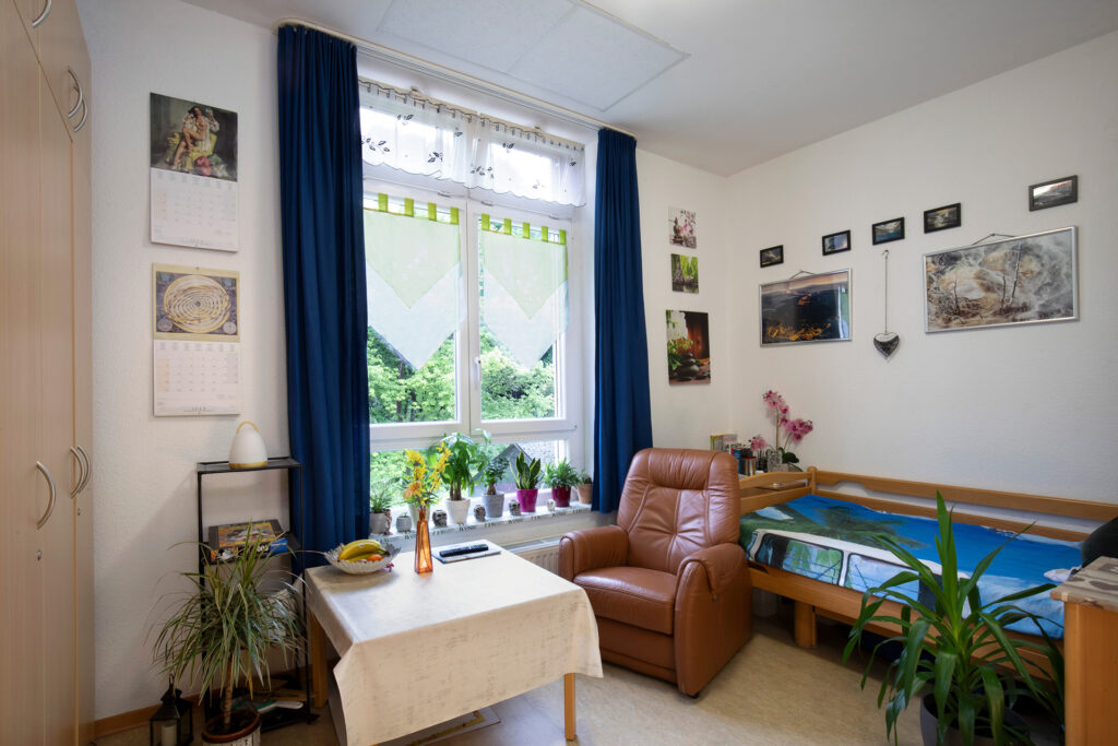 Blick in ein Bewohnerzimmer: Ein Fernsehsessel und ein Tisch stehen am Fenster, an der Wand steht ein Bett.