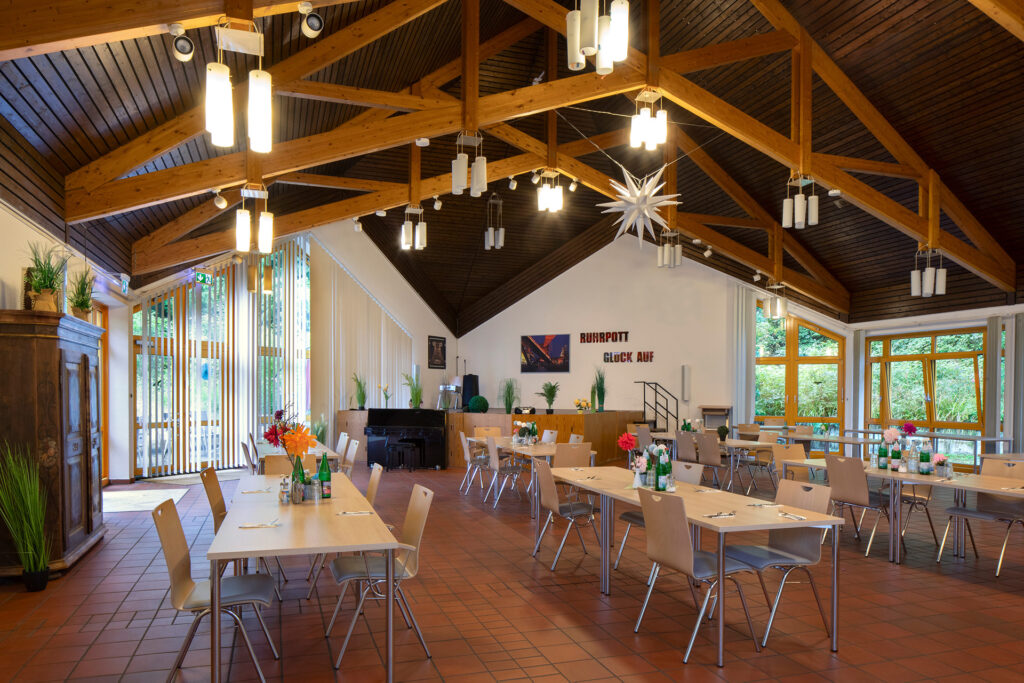 Blick in den Veranstaltungssaal: An zahlreichen Tischgruppen können viele Menschen Platz finden. Die erhöhte Bühne ist für alle gut sichtbar, davor steht ein Klavier. Bodentiefe Fenster und die hohe Decke sorgen für Helligkeit, ein antiker Schrank für Gemütlichkeit.