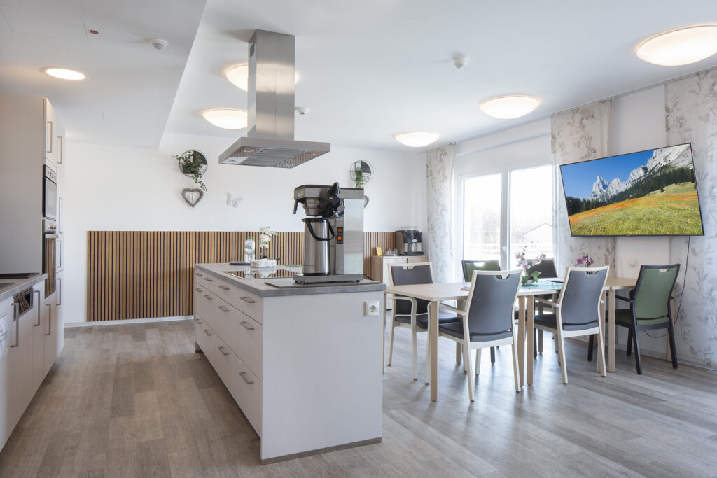 Blick in eine geräumige Gemeinschaftsküche: Neben einer Küchenzeile gibt es eine Kochinsel mit Theke und Sitzgruppen mit Tischen und Stühlen. Bodentiefe Fenster und Türen führen auf einen Balkon.