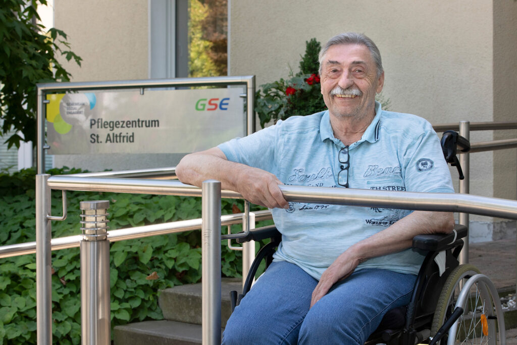 Ein Bewohner lächelt vor dem Schild des Pflegezentrum St. Altfrid in die Kamera