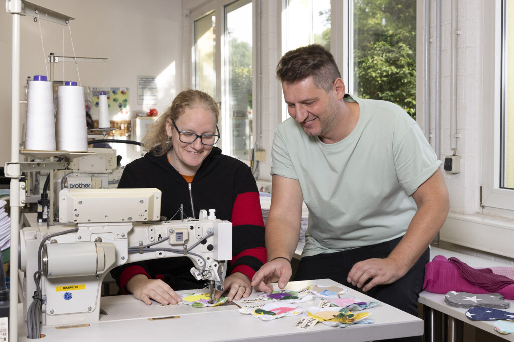 In der Näherei der GSE-Werkstatt Borbeck bespricht ein Gruppenleiter mit einer Werkstattmitarbeiterin ein Nähprojekt an der Nähmaschine.