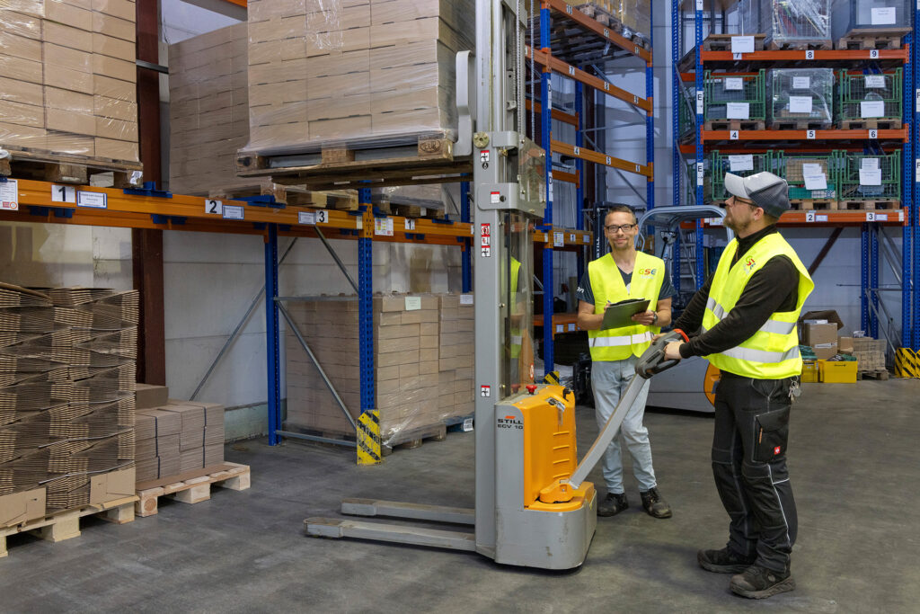 Im Lager fahren zwei Werkstattmitarbeiter einen Palettenhubwagen an das Palettenregal.